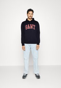 Felpa navy con "GANT" in rosso sul petto, abbinata a jeans azzurri chiari e sneaker grigie. Design semplice con una vestibilità comoda.