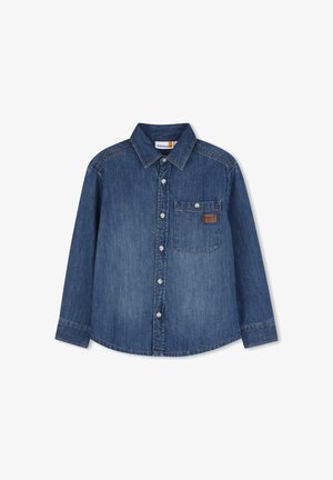 Chemise en denim bleu à manches longues avec boutons sur le devant, une poche poitrine unique avec fermeture par bouton, et une petite étiquette marron sur la poche.