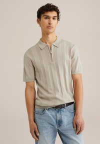 Polo de color beige claro con mangas cortas, que presenta una textura acanalada vertical y cierre de cremallera. Combinado con jeans de color azul claro y un cinturón marrón.