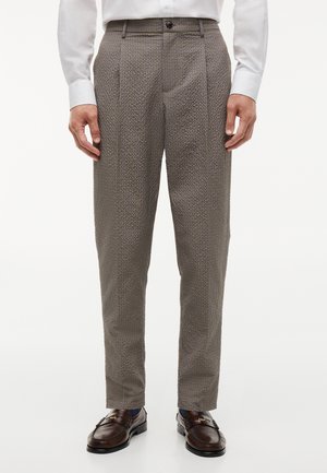 Man wearing slim-fit brown checkered trousers with a white shirt and dark brown leather loafers standing on a white background.