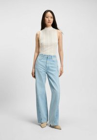 Kobieta z długimi ciemnymi włosami, ubrana w bezrękawnikową białą koronkową bluzkę, jasnoniebieskie dżinsy z wysokim stanem i szerokimi nogawkami oraz beżowe buty w szpic.