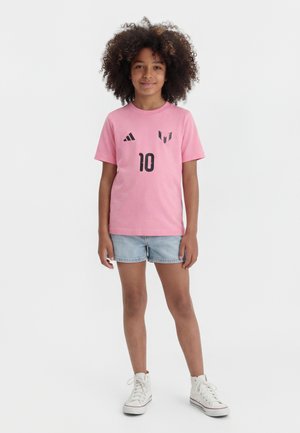 Παιδί με σγουρά μαλλιά, φορώντας ροζ αθλητικό μπλουζάκι με τον αριθμό 10, τζιν σορτς και λευκά ψηλά αθλητικά παπούτσια, στέκεται σε λευκό φόντο.