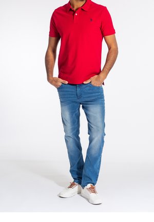 Homme debout les mains dans les poches, portant un polo rouge, un jean bleu et des baskets blanches avec des lacets marron sur un fond blanc uni.