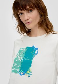 Sweatshirt blanc en coton avec un design graphique vibrante bleu et vert, incluant un élément floral et du texte disant "Passion". Texture lisse.