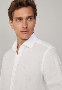 White linen shirt with a button-down collar, featuring a small blue embroidered emblem on the chest and subtle texture.