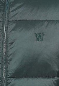 Nahaufnahme einer dunkelgrünen, gepolsterten Jacke mit sichtbarem Reißverschluss und gesticktem umgekehrtem "W"-Logo auf dem Gewebebereich.
