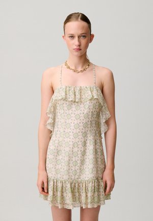 Giovane donna che indossa un abito senza maniche, arricciato, con fantasia, con sottili spalline e una collana a catena d'oro, in piedi davanti a uno sfondo uniforme.