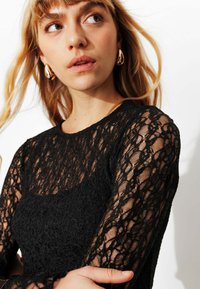 Haut en dentelle noire à manches longues, motif texturé et encolure ronde. Mis en valeur par un design transparent et une silhouette ajustée.