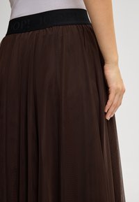 Mulher vestindo uma saia de malha castanho-escura com uma cintura elástica preta rotulada "JOOP!" e uma blusa clara por dentro, com a mão descansando ao lado.