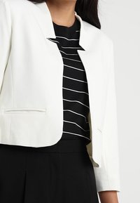Bílý přizpůsobený blazer nošený přes černobílý pruhovaný vršek, doplněný černými kalhotami s vysokým pasem.