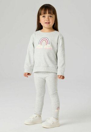 Nuori tyttö, jolla on päällään vaaleanharmaa pusero ja legginssit, joissa on sateenkaari- ja pilvikuvioita, seisoo yksinkertaisella vaalealla taustalla.