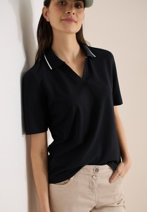 Femme portant un polo noir à manches courtes avec un col blanc bordé, et un pantalon beige, appuyée contre un mur de couleur claire.
