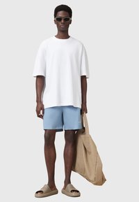 Camiseta blanca de gran tamaño, pantalones cortos azul claro, sandalias marrones de tipo slide en una persona que sostiene una bolsa de mano color beige. Diseño minimalista con una marca sutil.