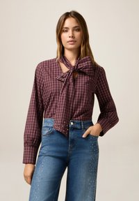 Bordeaux geruite blouse met strikdetail, lange mouwen. Gecombineerd met hooggeknoopte blauwe jeans met parelversieringen langs de zijkanten.