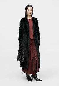 Černý kabát z umělé kožešiny, dlouhý a otevřený vpředu. Nošený přes vínové šaty s překládaným detailem. Černé kožené rukavice a černá texturovaná taška.