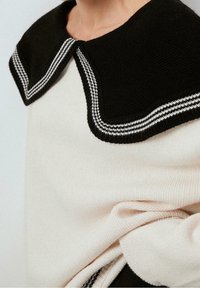 Strickpullover mit schwarzem, rundem Kragen mit weißen Streifen, cremefarbenem Körper und strukturiertem Stoff. Weiche, lockere Passform mit sichtbaren Nähten.