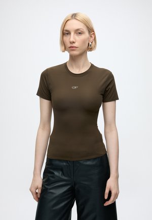 Ung kvinde med kort blondt hår iført en tætsiddende brun t-shirt og sorte læderbukser, stående mod en ensartet lys baggrund.