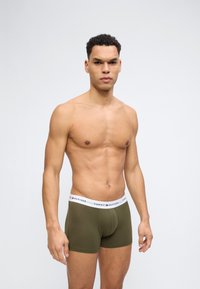 Άνδρας με κοντά σγουρά μαλλιά, φορώντας εσώρουχο μποξέρ Tommy Hilfiger σε ελιά πράσινο χρώμα με λευκή ζώνη μέσης με το λογότυπο, στέκεται μπροστά σε απλό ανοιχτό φόντο.