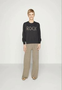 Černá mikina s dlouhým rukávem s nápisem "ROCK" z kamínků, spolu s béžovými širokými kalhotami. Modelka má na sobě zlaté boty a stojí před neutrálním pozadím.