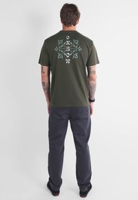 Camiseta de algodón verde oliva con un gráfico floral en blanco y teal en la parte posterior; combinada con pantalones gris oscuro. Presenta mangas cortas y cuello redondo.