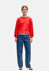 Roter Baumwollpullover mit langen puffärmel über einem weißen Rundhals, kombiniert mit lockeren blauen Jeans. Modell trägt schwarze Flip-Flops.