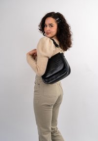 Black handbag in a textured crocodile pattern with a gold hardware accent. Worn crossbody style, paired with a beige sweater and light pants.