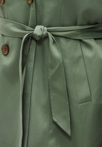 Πράσινο ελαιώδες παλτό τύπου τρεντσκότ δεμένο στη μέση με υφασμάτινη ζώνη και διαθέτει μεγάλα καφέ κουμπιά και πλαϊνή τσέπη.