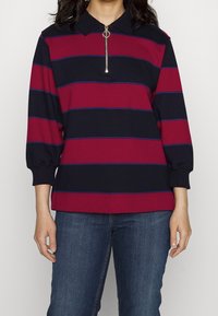 Tröja med långa ärmar och ett svart och vinrött randigt mönster, med dragkedja vid kragen och ribbstickade muddar, kombinerad med blå jeans i denim.