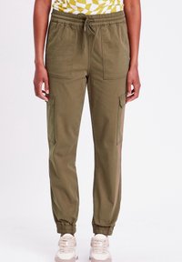 Pantalon de jogging vert olive avec une taille élastique, un cordon de serrage et des poches latérales. Ourlets effilés aux chevilles, fabriqué en tissu doux.