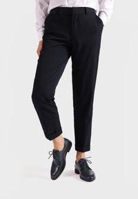 Pantaloni neri su misura con risvolti arrotolati, abbinati a scarpe in pelle scura con punte arrotondate e dettagli con lacci. Texture liscia nel complesso.