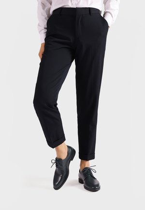 Pantaloni neri su misura con risvolti arrotolati, abbinati a scarpe in pelle scura con punte arrotondate e dettagli con lacci. Texture liscia nel complesso.