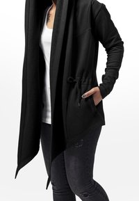 Cárdigan negro con capucha y apertura frontal, cinturilla ajustable con cordón y bolsillos; confeccionado en una tela suave con una textura lisa.