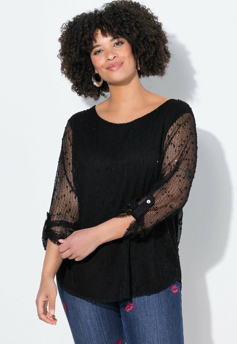 Top nero con una vestibilità ampia, caratterizzato da maniche trasparenti con perline e bottoni neri ai polsini. Abbinato a jeans blu con stampa di labbra.