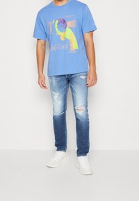 Hombre vistiendo una camiseta gráfica azul, jeans desgastados y rasgados, y zapatillas blancas, de pie contra un fondo claro. 