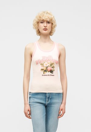 Femme aux cheveux blonds bouclés portant un débardeur rose avec un motif de fraise et du texte, associée à un jean bleu clair, debout sur un fond blanc.