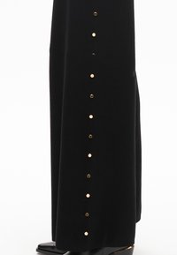 Černá sametová sukně s pozlacenými knoflíkovými akcenty po straně. Tkanina má jemnou strukturu, která elegantně splývá až k podlaze.