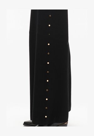 Falda de terciopelo negro con acentos de botones dorados a lo largo del lado. La tela tiene una textura suave, cayendo elegantemente hasta el suelo.
