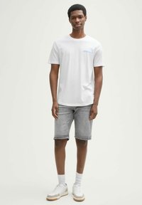 Weißes T-Shirt mit kurzen Ärmeln und einer blauen Akzenttasche, kombiniert mit grauen Denim-Shorts und weißen Sneakers mit sandfarbenen Akzenten.