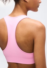 Γυναίκα που φαίνεται από πίσω φοράει αθλητικό σουτιέν ροζ ανοιχτό χρώμα με σχέδιο racerback, σε απλό ανοιχτό φόντο.