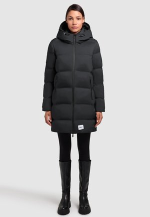 Zwarte pufferjas met capuchon, gewatteerd ontwerp, rits aan de voorkant en zijvakken. Draagt over zwarte leggings en knielange zwarte laarzen.