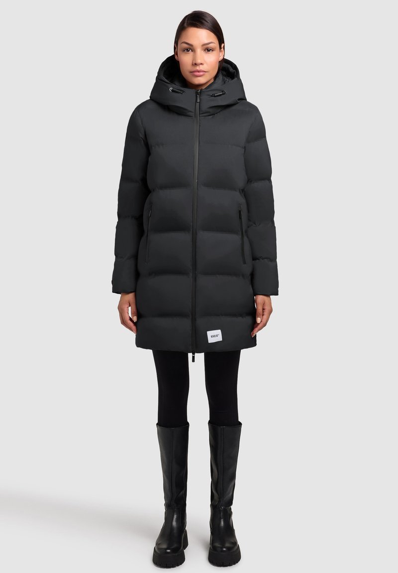 Black puffer coat with a hood, quilted design, front zipper, and side pockets. Worn over black leggings and knee-high black boots.
