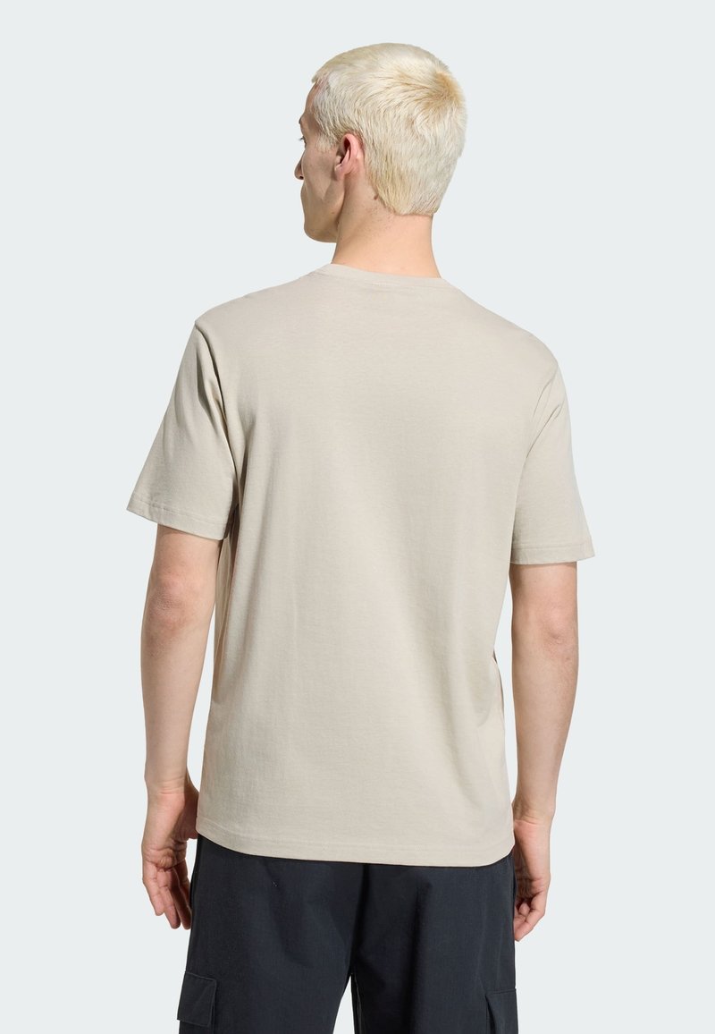 Man met kort blond haar, die een effen beige T-shirt met korte mouwen en een zwarte broek draagt, staat met zijn rug naar voren gericht.