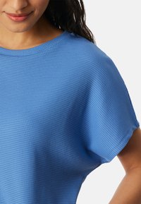 Frau trägt ein strukturiertes, leuchtend blaues Kurzarmoberteil mit rundem Ausschnitt, dargestellt vom Hals bis zur Mitte des Oberkörpers.