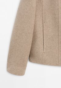 Close-up of a beige wool coat sleeve and side, showing a vertical slit pocket and soft textured fabric.