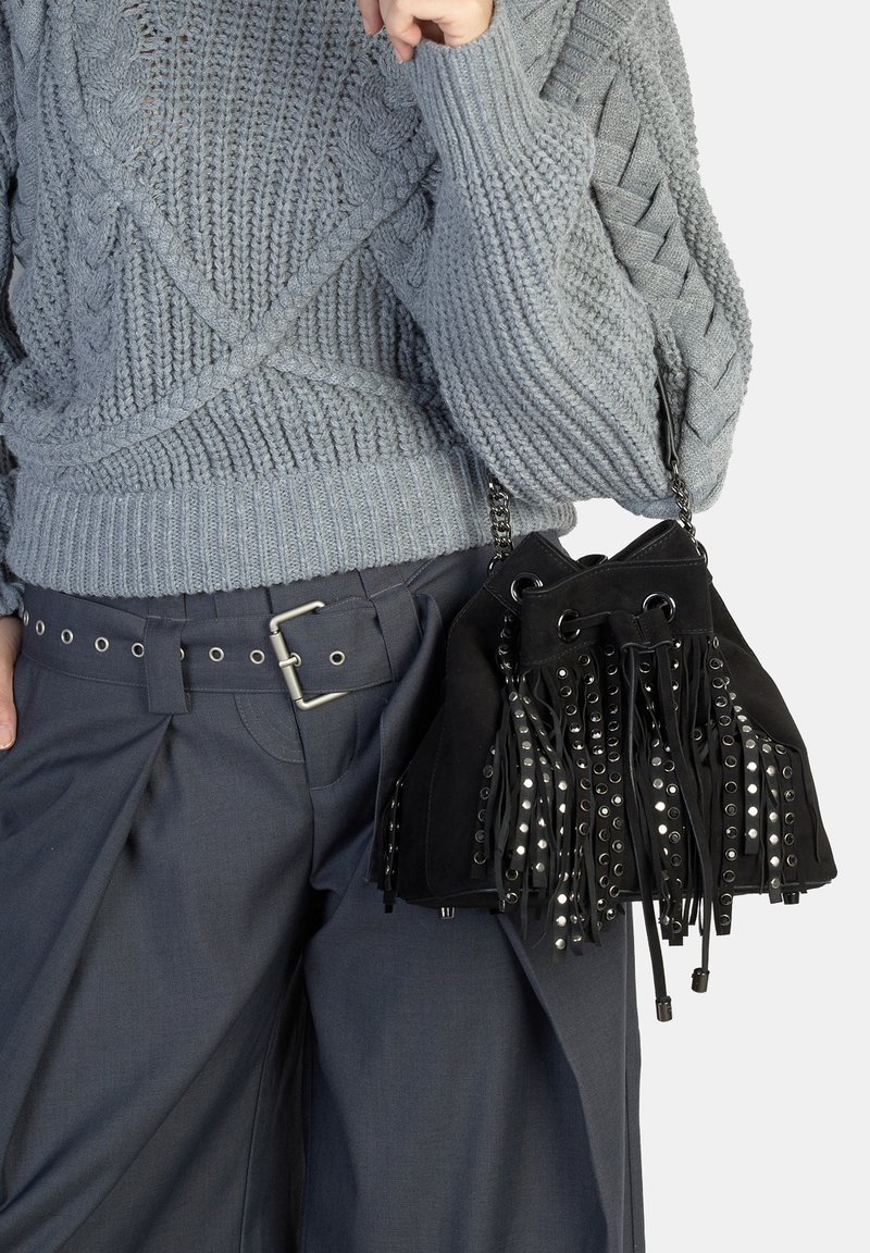 Czarna torebka zamszowa z frędzlami i srebrnymi oczkami. Połączona z szarym swetrem warkoczowym i ciemnymi spodniami z paskiem na klamrę.