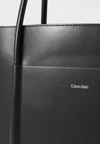 Borsa tote in pelle nera con una superficie liscia, dotata di una tasca frontale e un'etichetta in rilievo "Calvin Klein" in bianco. Design minimalista.