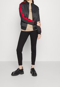 Svart bomberjacka med strukturerat mönster och röda sidostrick samt en beige topp och svarta åtsittande byxor. Chunky svarta skor fulländar looken.