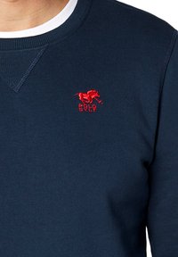Granatowy sweatshirt z prążkowanym dekoltem, teksturowanym materiałem i czerwonym wyszywanym logo konia z napisem "POLO SYLT" poniżej.