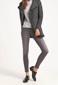 Abrigo gris texturizado con solapas grandes, combinado con un suéter gris claro con adornos y vaqueros ajustados gris oscuro. Mocasines negros.