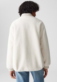 Witte, donzige fleece jas met een hoge kraag, gedropte schouders en ribgebreide manchetten. Zachte textuur, relaxte pasvorm en geen zichtbare sluitingen.